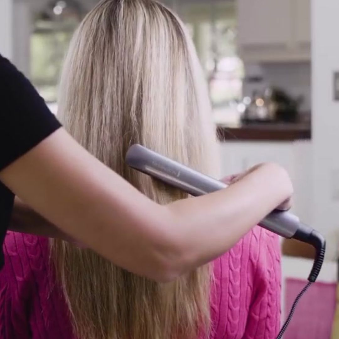 Person using a hair straightener on another person's hair in a kitchen setting