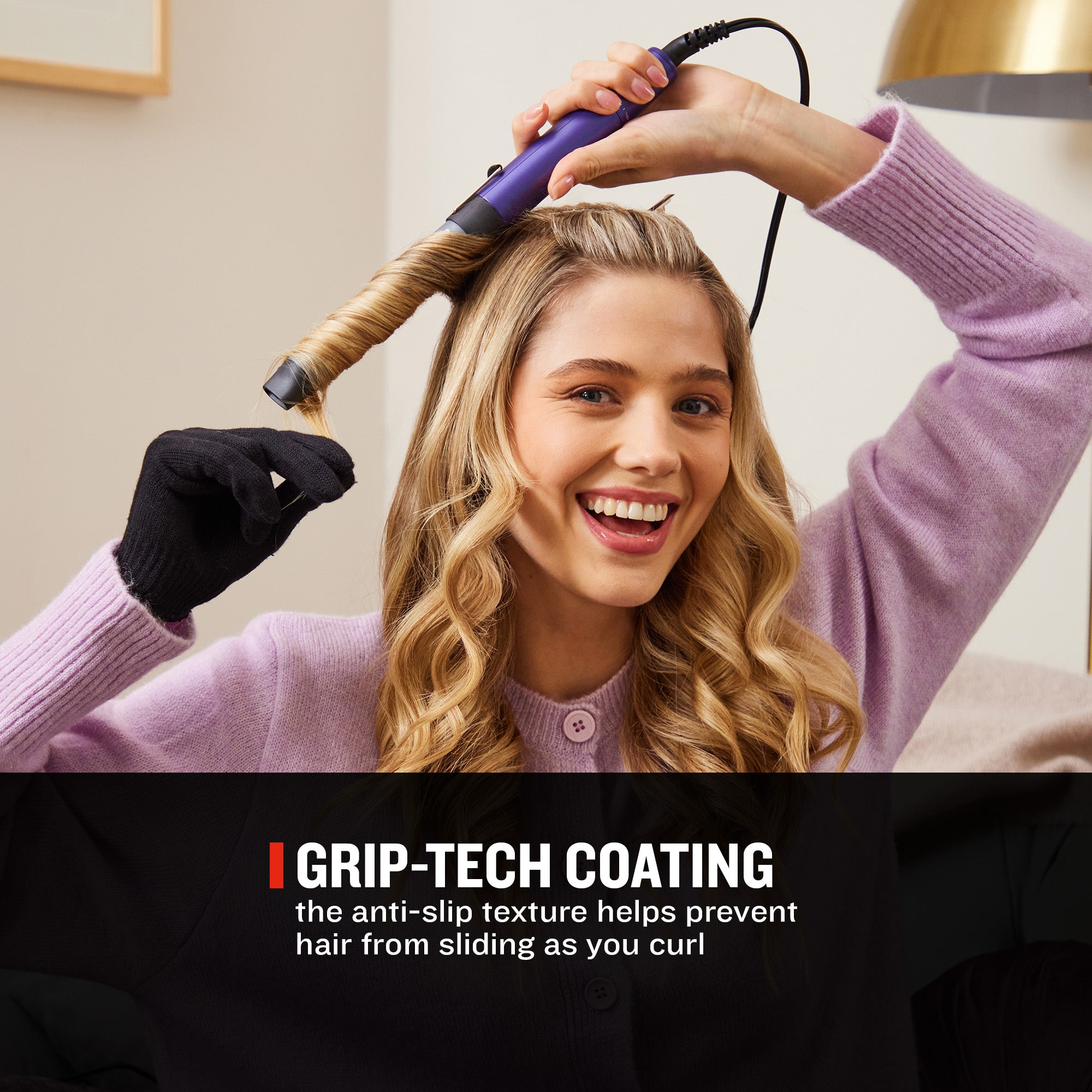 A woman with long blonde hair uses a purple and black curling iron to curl a section of her hair. She is wearing a lavender knit sweater and a black glove.