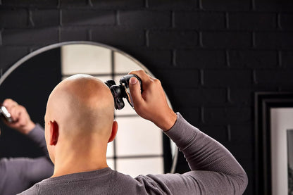 Person using a hair trimmer on their head in front of a mirror with a black brick wall background.