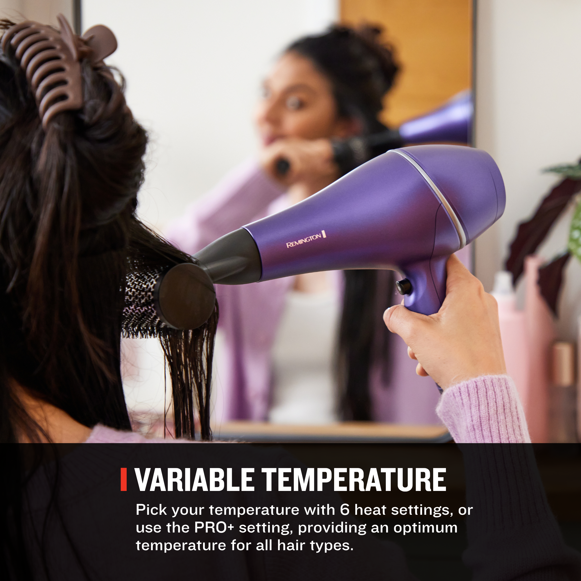 A person using a purple hair dryer on their hair, with a mirror in the background showing the person's face. A text banner at the bottom reads 'VARIABLE TEMPERATURE'.
