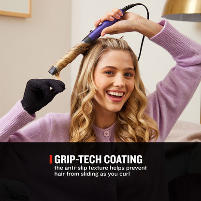 A woman with long blonde hair uses a purple and black curling iron to curl a section of her hair. She is wearing a lavender knit sweater and a black glove.