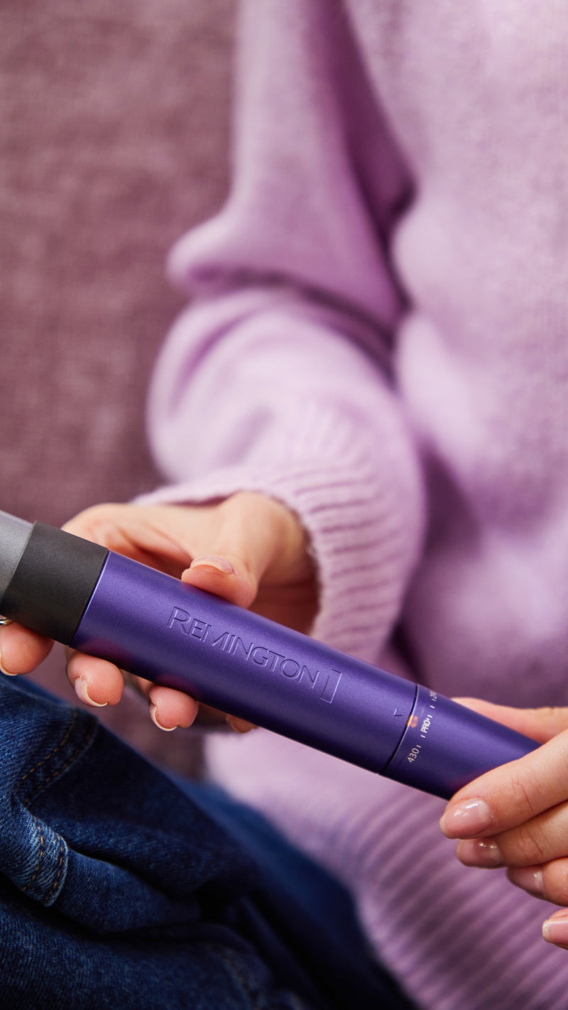 woman holding a purple Remington curling wand, wearing a lavender jumper and blue jeans