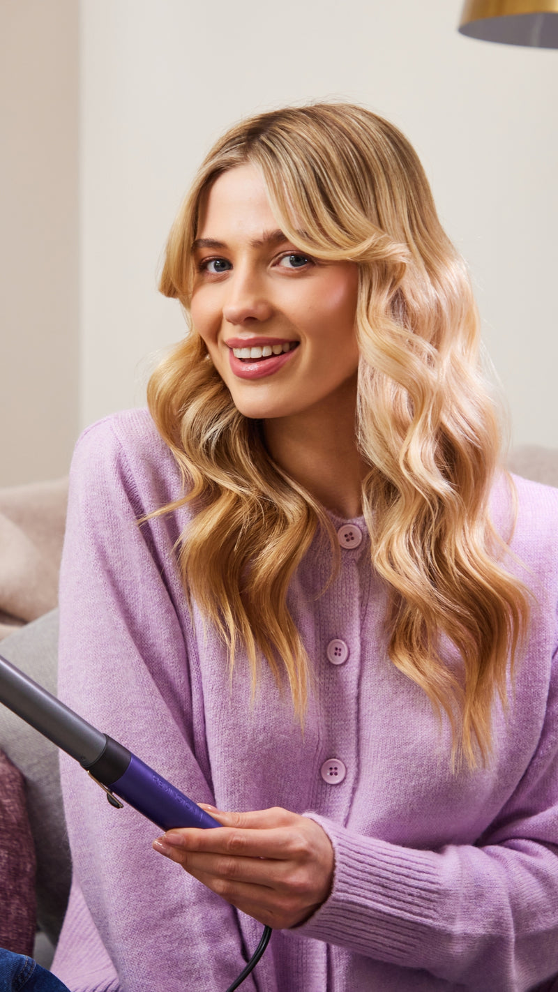 A smiling blonde woman with wavy hair wears a lavender cardigan and holds a purple and black hair styling tool in a cozy indoor setting.