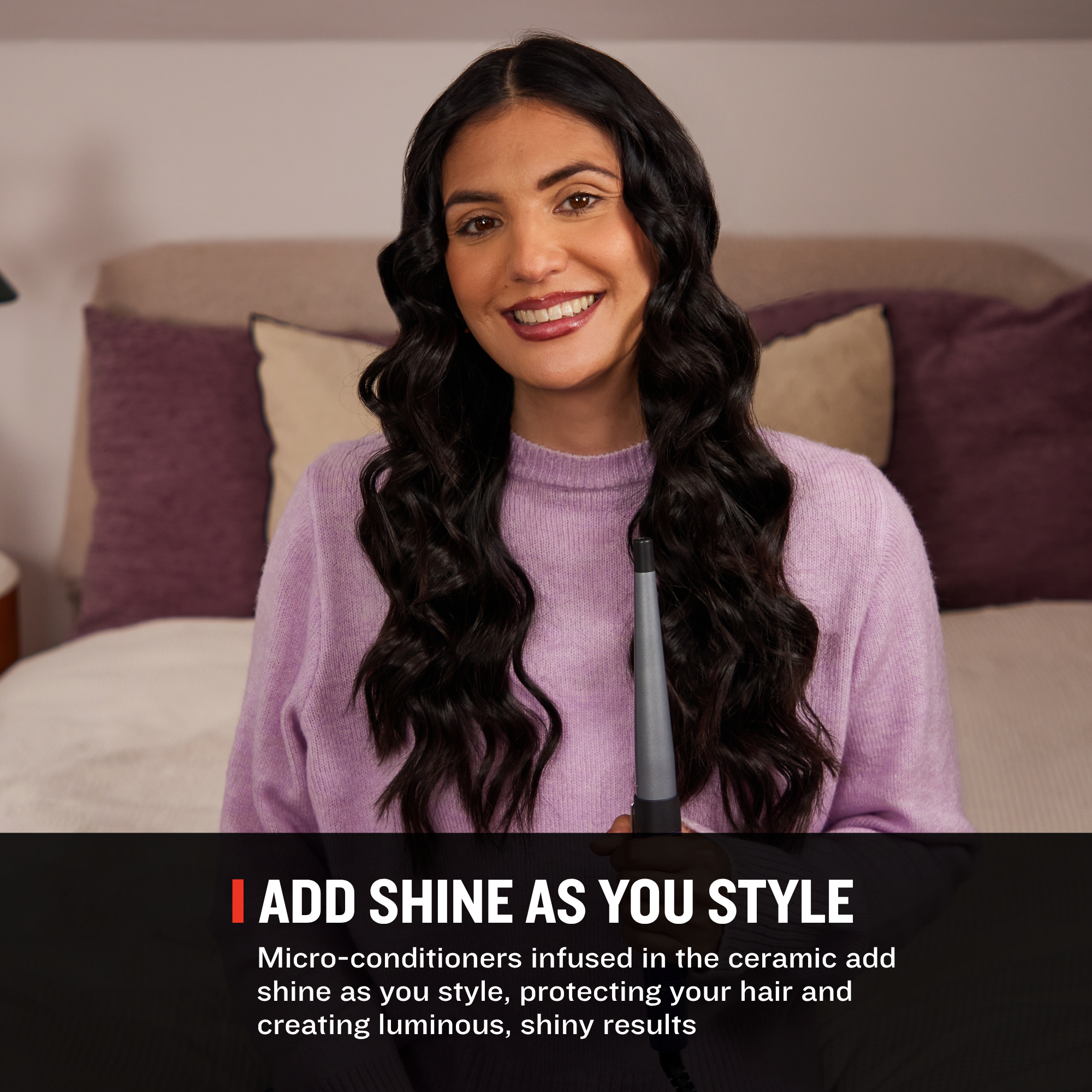 A smiling woman with long, dark, glossy wavy hair wearing a lavender sweater, holding a hair styling tool in front of a bed with purple and beige pillows. A black banner at the bottom reads 'ADD SHINE AS YOU STYLE' with descriptive text about micro-conditioners and ceramic shine technology.