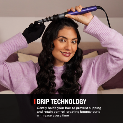 A woman with long dark wavy hair uses a purple curling iron on her hair in a bedroom setting. She is wearing a light purple sweater and a black glove.