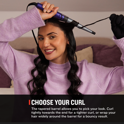 A woman with long dark wavy hair uses a purple and silver curling iron on her hair in a bedroom setting. A black text banner at the bottom reads 'CHOOSE YOUR CURL' with descriptive text about curling hair.