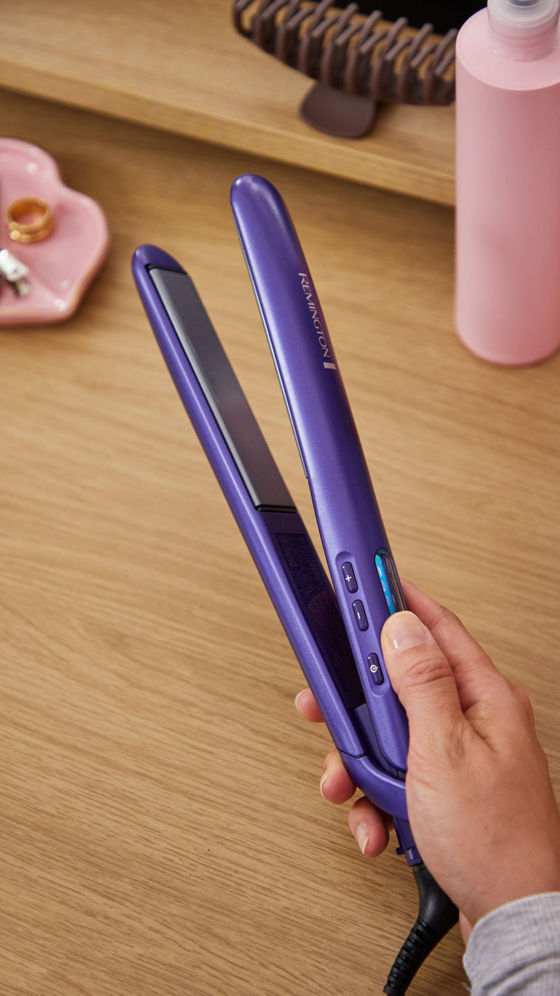 A hand holding a purple hair straightener on a wooden desk, with a pink bottle and other desk items blurred in the background.