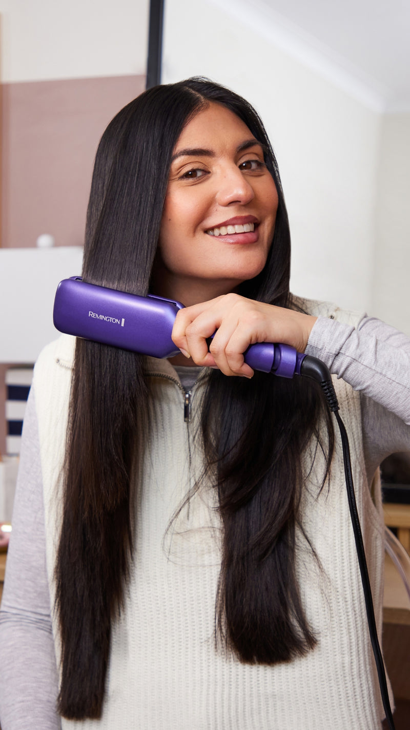 A woman with long, straight dark hair uses a purple hair straightener on her hair in an indoor setting.