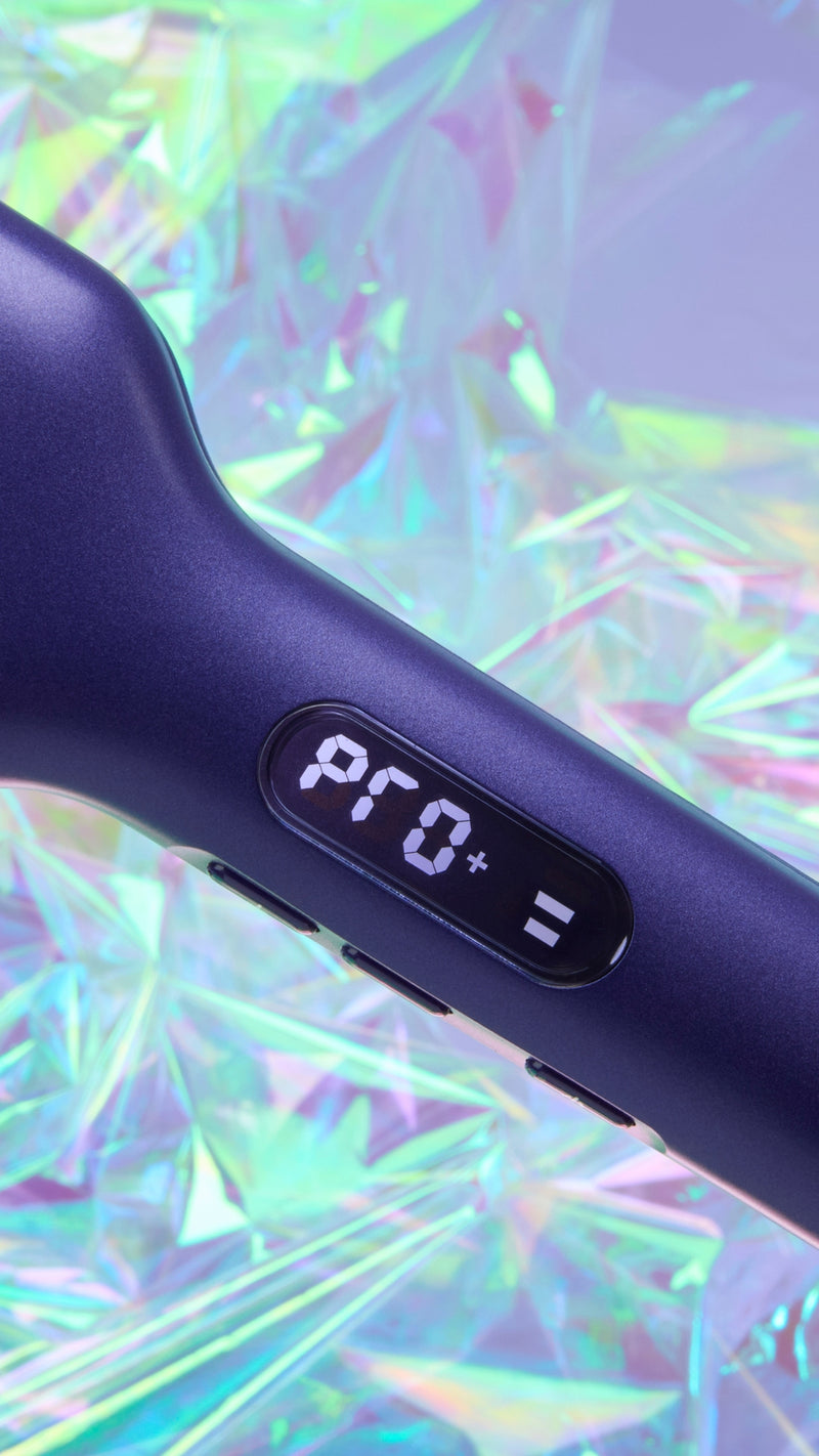 Close-up of a dark purple hair straightener showing the 'PRO+' setting on its control panel, set against a vibrant, iridescent holographic background.