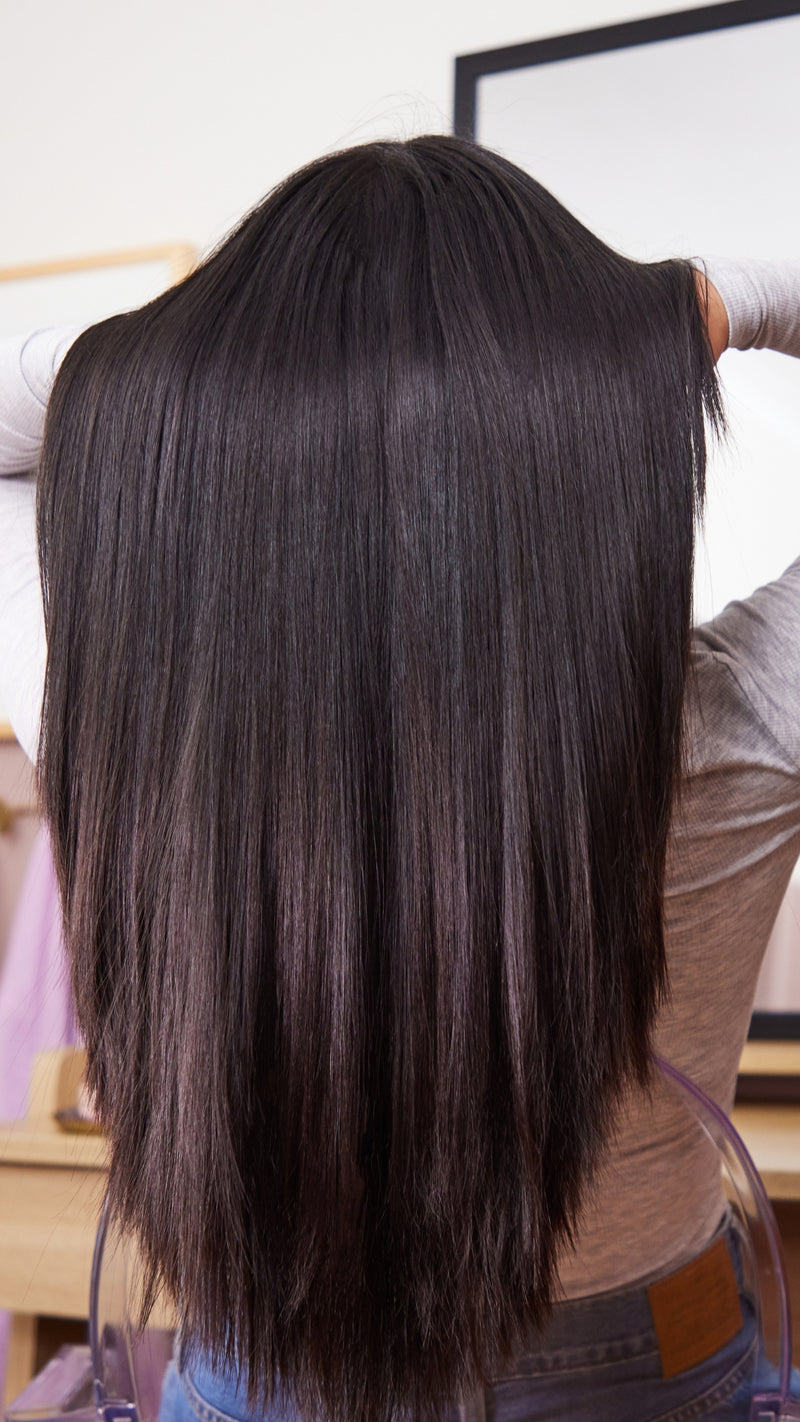A person viewed from behind with very long, straight, dark brown to near-black hair, wearing a light gray top and blue jeans in an indoor setting.