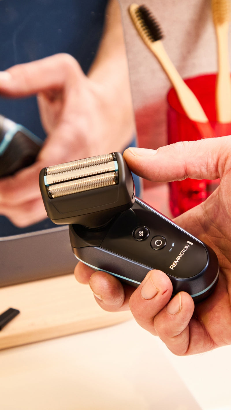 A close-up of a person holding a black and silver electric shaver in their hand, with a bathroom mirror reflection and a toothbrush holder in the background.