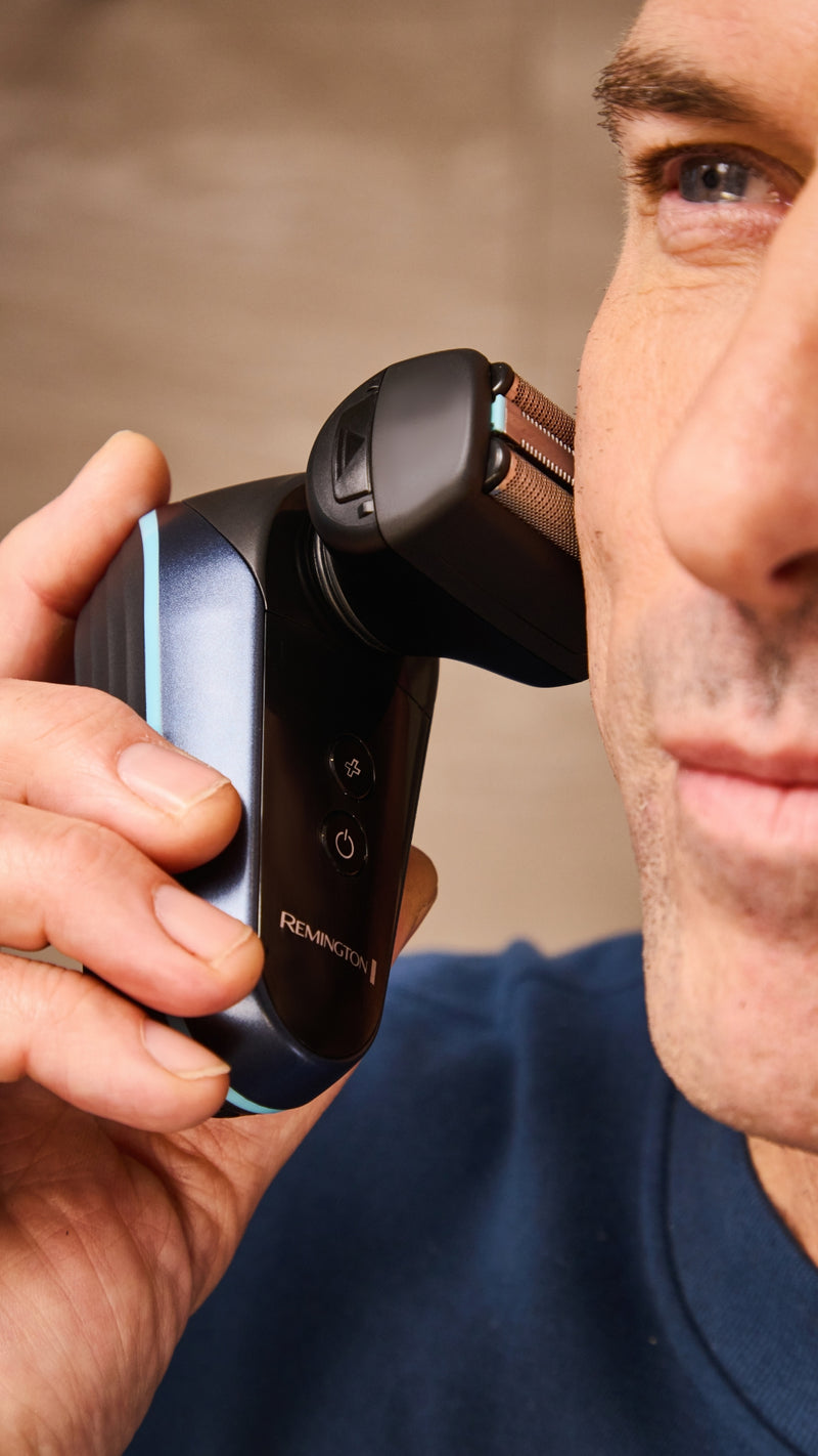 A close-up lifestyle shot of a man using a black and blue Remington Electric Foil Shaver on his face, showing the product in use.