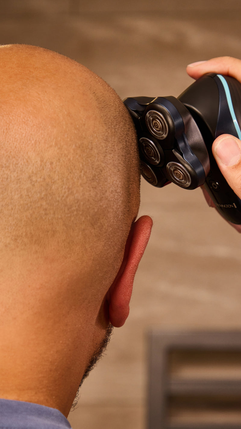 A close-up lifestyle shot of a person shaving their head with a black electric shaver with a blue accent line.