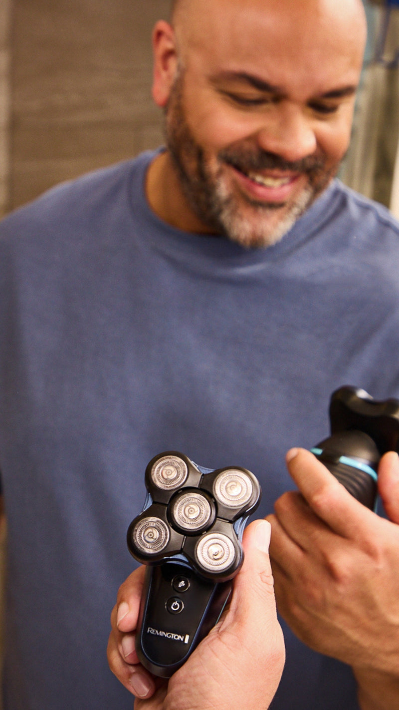 A smiling man in a blue shirt holding an  electric shavers, clearly showing the five rotating blade heads of a Remington Balder in a mirror reflection