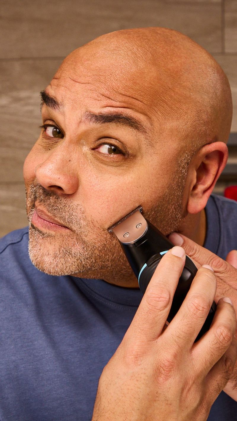 A close-up of a man using an electric trimmer on his facial hair, wearing a blue t-shirt against a neutral background.